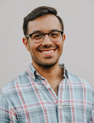 The image shows a man wearing glasses, smiling at the camera, with dark hair and a beard. He is dressed in a plaid shirt and has a light-colored background behind him.