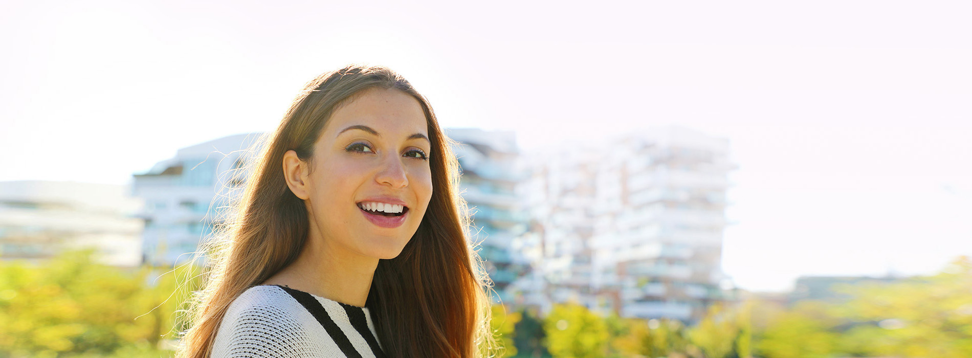The image is a photograph of a young woman with long hair smiling at the camera, wearing a black and white striped top, against a blurred background that suggests an outdoor setting.