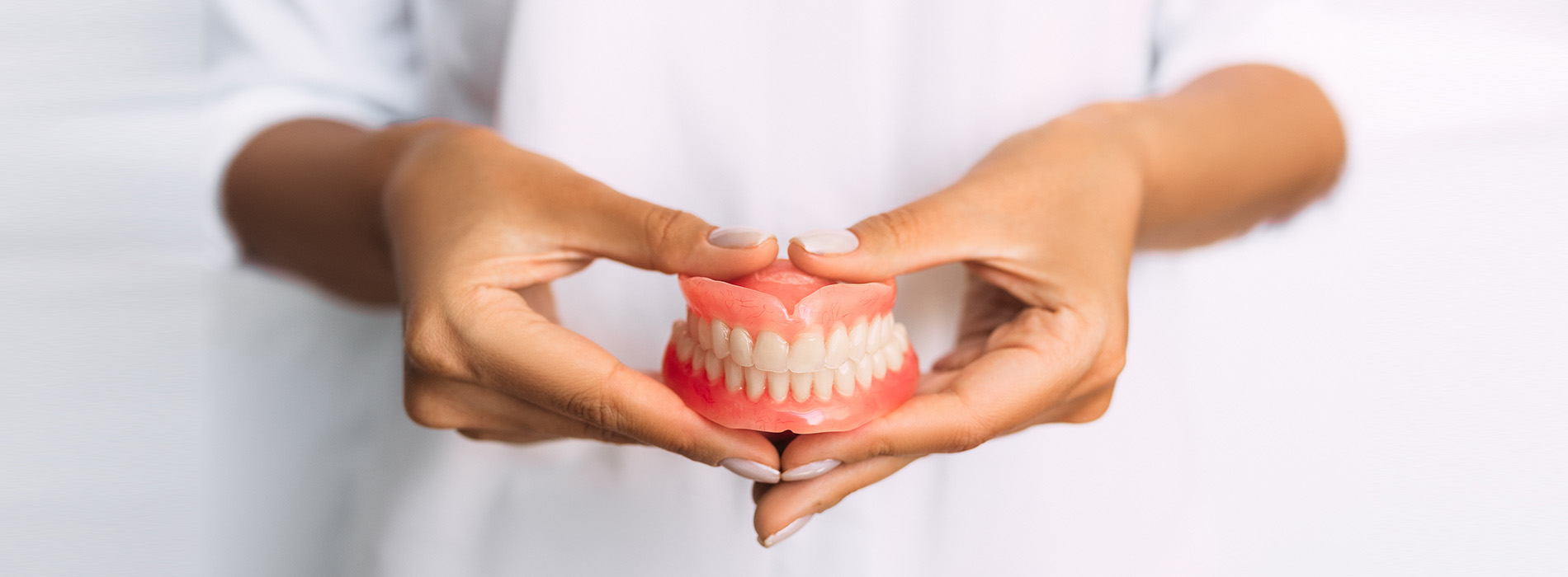 The image shows a person s hands holding a pair of dentures, with the focus on the dental appliance, set against a blurred background.