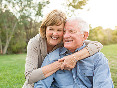 An elderly couple embracing, with the woman wearing a white top and the man in a blue shirt, both smiling and looking at each other against a backdrop of greenery.