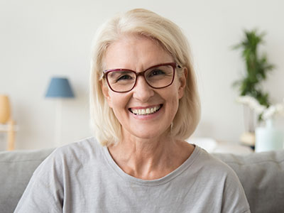 The image shows a woman with short blonde hair, wearing glasses and a gray top, smiling at the camera. She is seated indoors, possibly in a living room, with a neutral-toned background that includes a lamp and a framed picture.