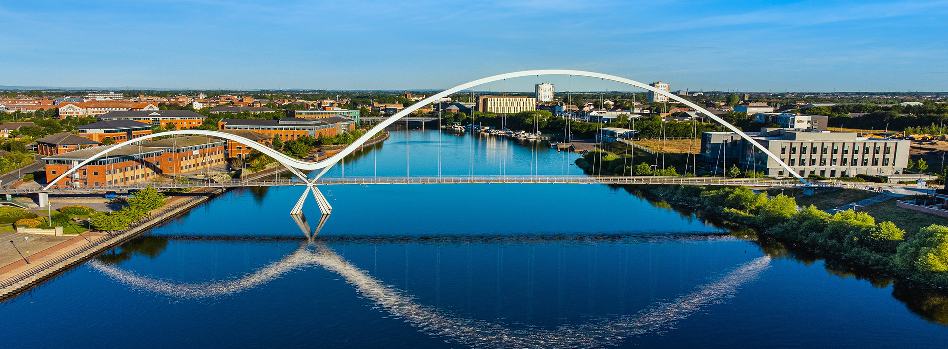 The image is a photograph of a modern, architectural pedestrian bridge spanning over a calm body of water, with the city skyline in the background and a clear blue sky above.