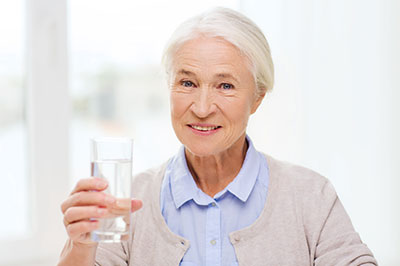 The image shows an elderly woman in a domestic setting, holding a glass of water and smiling at the camera.