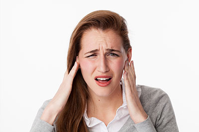 The image shows a young woman with her hand on her head, appearing to be in pain or discomfort, with an expression of distress. She is wearing a gray cardigan and has short brown hair.