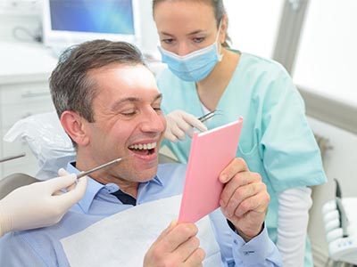 The image depicts a dental office setting where a man is sitting in the dentist s chair, smiling broadly at a pink card he holds. A female dental professional stands behind him, looking on with a smile.