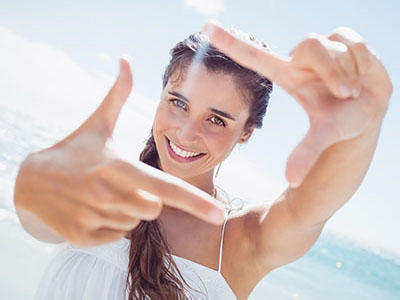 The image shows a woman holding up her index finger in front of her face, smiling, with a clear blue sky and ocean behind her.